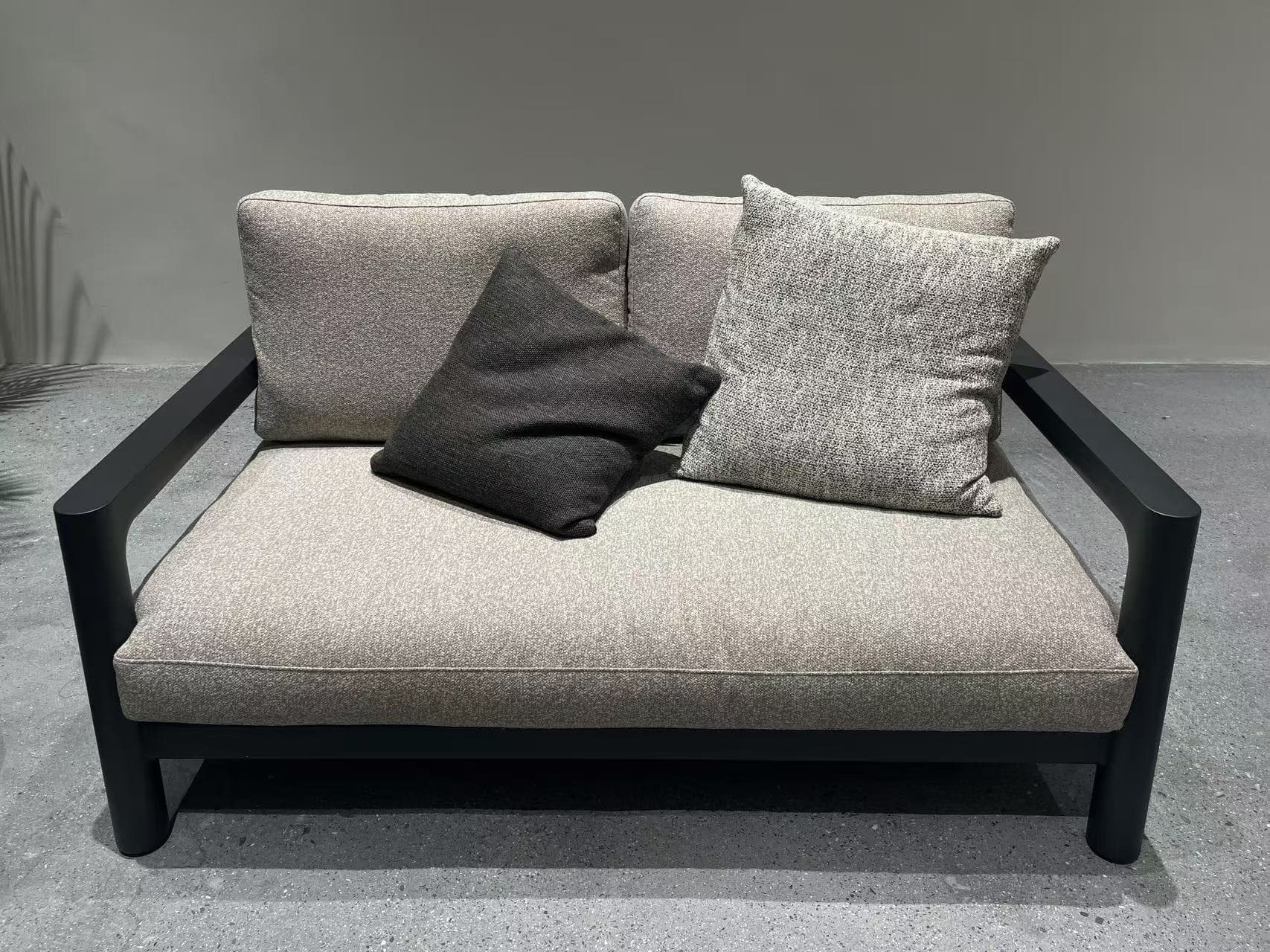 A light gray outdoor sofa with two light gray throw pillows and two dark brown throw pillows is placed on a light gray textured rug in a modern setting.